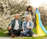 Auf dem Bild sind eine Frau und ein älterer Mann zu sehen. Sie sitzen auf dem Boden und schauen sich an. Im Hintergrund spielt ein Mädchen mit einer Flagge der Ukraine.