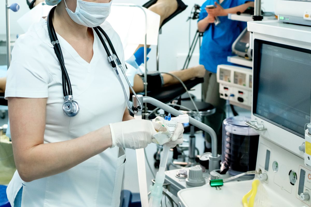Eine Ärztin mit Stethoskop steht vor einem Monitor und hält einen Beatmungsschlauch in der Hand. Im Hintergrund liegt ein Patient auf einer Liege und ist an ein Blutdruck-Messgerät angelegt. Daneben steht eine Krankenschwester.