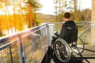 Ein junger Erwachsener im Rollstuhl auf einem Balkon sieht auf einen Waldsee. Ihm strahlt die untergehende Sonne entgegen.