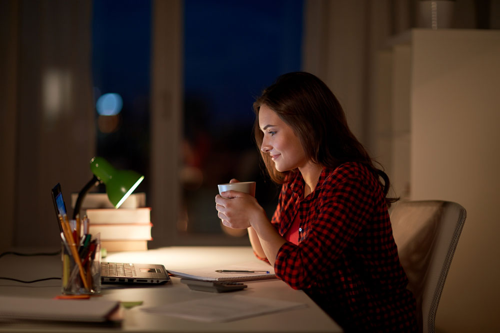 Frau mit Becher in der Hand sitzt am Abend vor dem Computer.