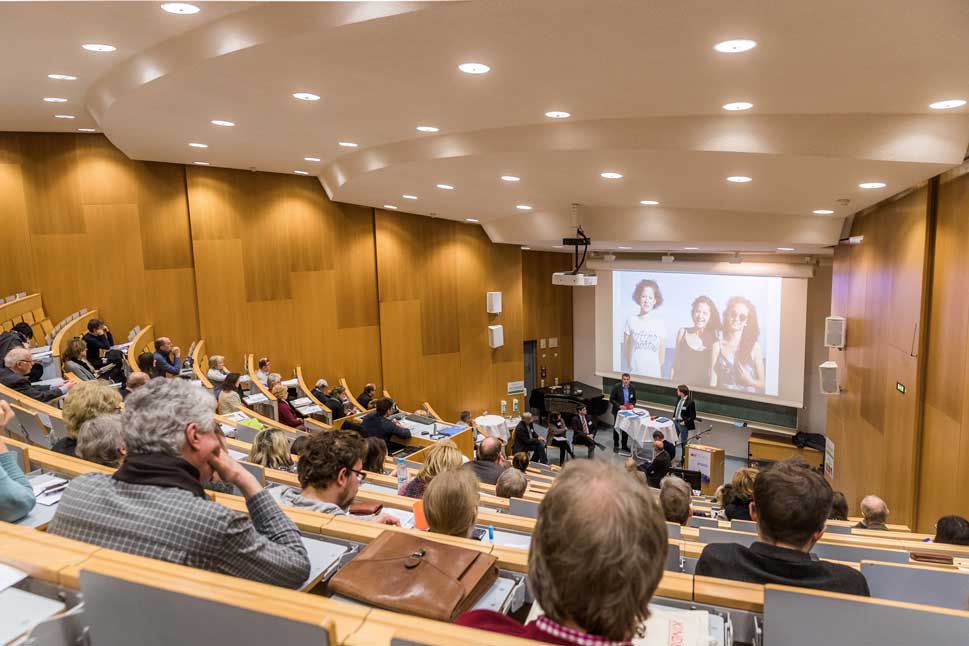 Viele Menschen in einem Hörsaal hören einer Rede zu.