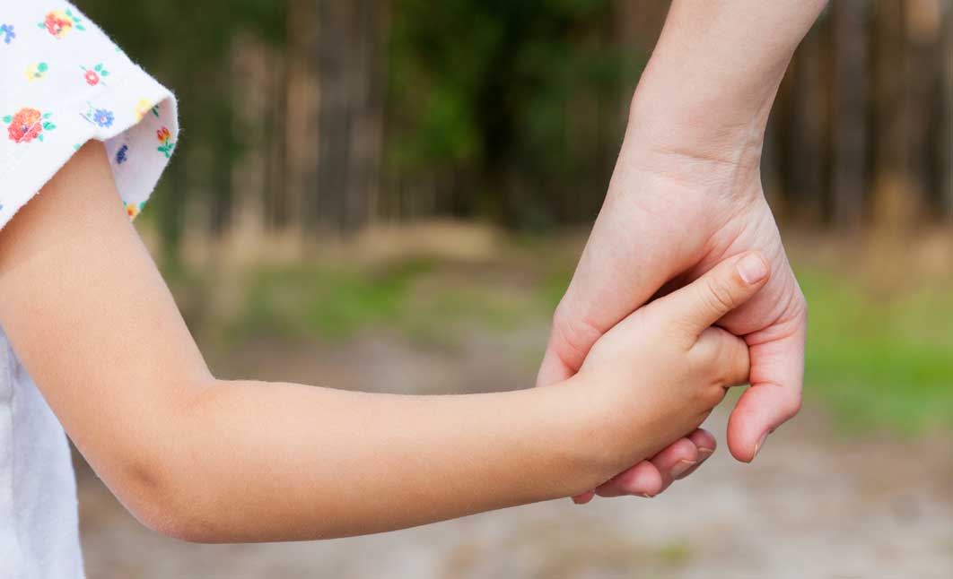Kostenloser Eintrag in unsere Elterndatenbank Eine Frauenhand hält eine Kinderhand. Im Hintergrund ist ein Waldgebiet unscharf dargestellt.